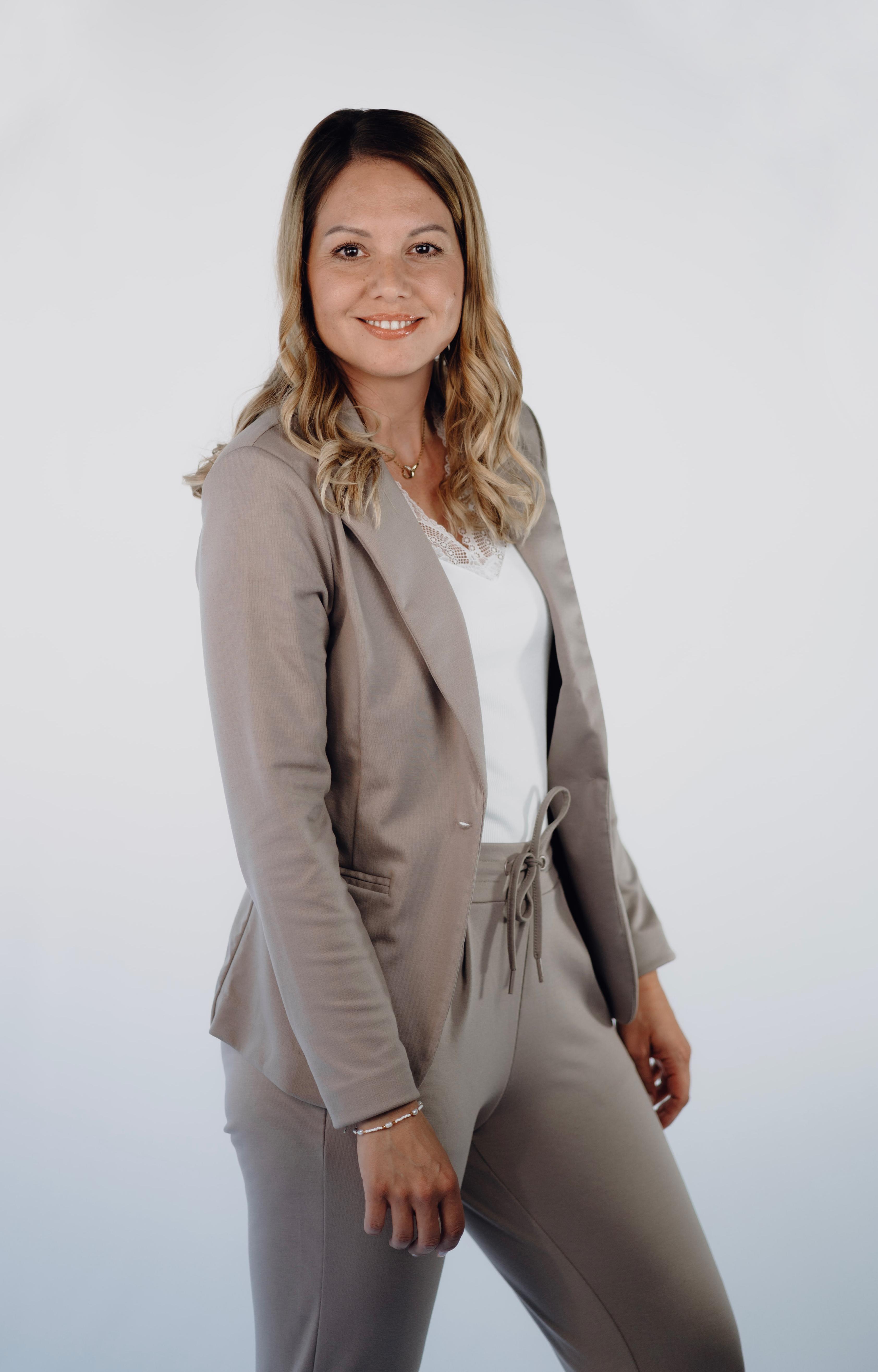 Professionelles Porträtfoto einer Frau mit gewelltem, blond meliertem Haar, lächelnd, in einem taupefarbenen Blazer mit passender Hose über einem weißen Top.